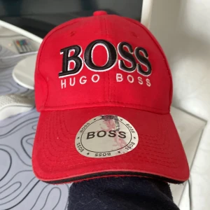 Röd keps från Hugo Boss - Snygg röd keps från Hugo Boss med stor broderad logotyp framtill. Perfekt för att ge en färgklick till din outfit. Justerbar passform för optimal komfort. Lappen på skärmen går/ska man ta bort men är så lite använd så har inte blivit av :)