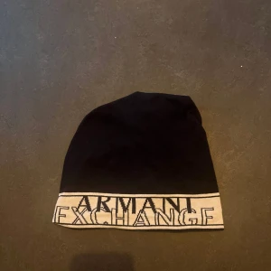 Svart marinblå mössa från Armani Exchange - Snygg svart marinblå  mössa från Armani Exchange med vit kant och logotyp. Perfekt för att hålla värmen med stil. Tillverkad i mjukt material för en bekväm passform.