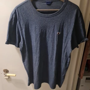 Blå t-shirt från Champion - Säljer en klassisk blå t-shirt från Champion. Den har en liten logga på bröstet och är perfekt för en avslappnad stil. T-shirten är kortärmad och gjord i ett mjukt material som känns skönt mot huden. Perfekt för vardagsbruk!