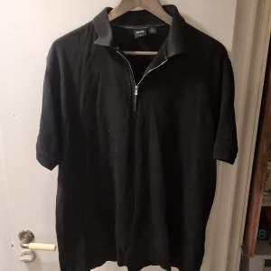 Svart pikétröja från Hugo Boss - Snygg svart pikétröja från Hugo Boss med dragkedja vid halsen. Perfekt för en stilren look. Tillverkad i Thailand.