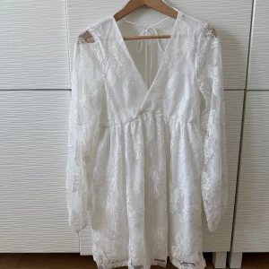 Vit blommig klänning med spetsdetaljer - Säljer en vacker vit klänning med blommigt spetsmönster och v-ringning. Klänningen har långa ärmar och en knytning i ryggen. Perfekt för sommarens festligheter!  Perfekt som studentklänning eller till midsommar. Innerklänning så man ej ser igenom. Köpte för att ha till studenten, men alldeles för kort för mig, därav prislapp kvar och aldrig använd. Lite skimrigt i tyget. Från GinaTricot, storlek S.