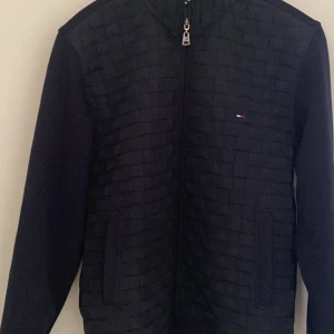 Svart quiltad jacka från Tommy Hilfiger - Snygg svart quiltad jacka från Tommy Hilfiger med dragkedja framtill. Jackan har ett diskret märke på bröstet och långa ärmar. Perfekt för en stilren look. Blå/svart i färgen.