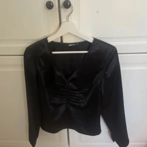 Svart glansig topp från Gina Tricot - Säljer en elegant svart glansig topp från Gina Tricot. Toppen har en v-ringning och långa ärmar, perfekt för en stilren look. Passar utmärkt till både fest och vardag. 🖤
