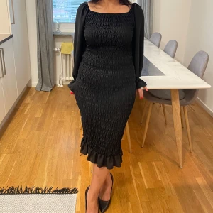 Svart off shoulder klänning med volangdetaljer - Snygg svart klänning med smockad design och volangdetaljer vid fållen. Klänningen har långa ärmar med elastiska manschetter och en elegant, figurnära passform. Perfekt för en stilren look. Storlek S-M, den är väldigt elastisk 