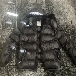 Moncler jacka - Säljer en snygg svart pufferjacka från Moncler. Jackan har en quiltad design med dragkedja och huva. Perfekt för kyliga dagar med sin stilrena look och varma fyllning.