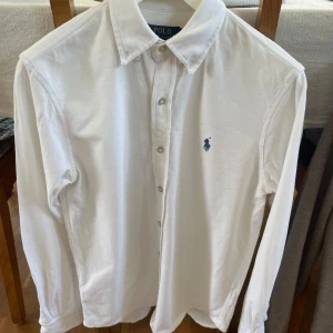 Vit skjorta från Ralph Lauren - Säljer en klassisk vit skjorta från Ralph Lauren med den ikoniska loggan broderad i blått på bröstet. Skjortan har långa ärmar och knappar framtill. Perfekt för en stilren look.