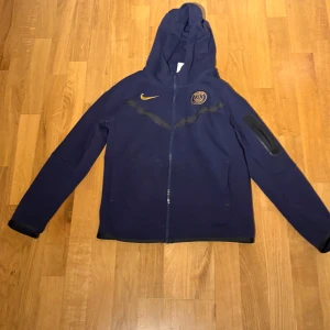 Mörkblå huvtröja från Nike tech psg  - Säljer en mörkblå huvtröja från Nike med dragkedja och PSG-logga på bröstet. Tröjan har långa ärmar och en huva, samt gula detaljer. Perfekt för fotbollsfans!