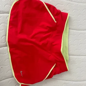 Golf/sportkjol från Nike - Snygg röd sportkjol från Nike med Dri-FIT-teknologi. Kjolen har en praktisk dragkedja baktill och kontrasterande gula detaljer. Perfekt för träning eller golf samt tennis. Inbyggda shorts för extra komfort.