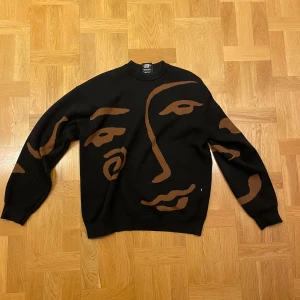 Snygg tröja från Sweet SKTBS - Säljer en unik svart tröja från Sweet SKTBS med ett konstnärligt ansiktsmönster i brunt. 