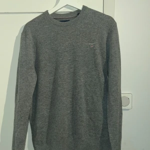 Grå stickad tröja från Gant - Säljer en stilren grå stickad tröja från Gant. Tröjan har en rund halsringning och långa ärmar. Perfekt för kyligare dagar och passar till många olika outfits. En liten logga pryder bröstet för en diskret touch av stil.