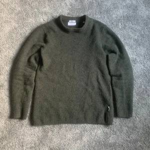 Grön ulltröja från Lindbergh - Säljer en grön tröja i lammull från Lindbergh. Tröjan har en avslappnad passform med ribbstickad krage, ärmslut och nederkant. Perfekt för kyliga dagar.