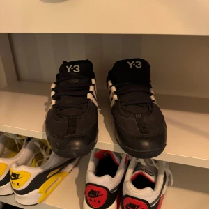 Svarta Y-3 sneakers - Snygga svarta Y-3 sneakers med vita detaljer och snörning. Skorna har en modern design med Y-3 logga på plösen och tre vita ränder på sidan. Perfekta för en stilren look.