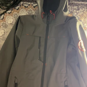 Grå jacka från Helly Hansen - Säljer en grå jacka från Helly Hansen med dragkedja och huva. Jackan har en patch på ärmen och orangea detaljer vid snörningen. Perfekt för utomhusaktiviteter. Pris kan diskuteras 