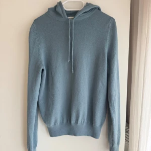 Ljusblå hoodie från Soft Goat - Säljer en supermjuk ljusblå hoodie från Soft Goat i 100% kashmir. Tröjan har en klassisk design med huva och dragsko, samt långa ärmar och ribbade muddar. Perfekt för en stilren och bekväm look.