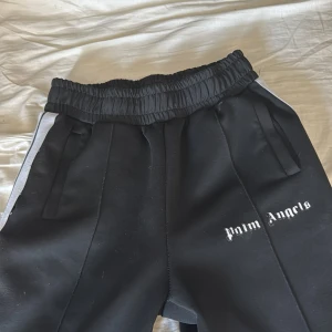 Svarta mjukisbyxor från Palm Angels - Snygga svarta mjukisbyxor från Palm Angels med vit logga på benet. Byxorna har elastisk midja och vita ränder längs sidorna. Perfekta för en avslappnad stil. Loggan är ganska sliten men annars i hyfsat skick