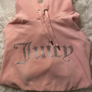 Rosa hoodie från Juicy Couture - Säljer en snygg rosa hoodie från Juicy Couture med glittrande stenar som bildar texten 'Juicy' på framsidan. Hoodien har långa ärmar och en justerbar huva med dragsko. Perfekt för en avslappnad stil.