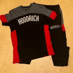 Röd hoodrich tracksuit - Säljer en cool t-shirt från Hoodrich i svart, grått och rött. T-shirten har korta ärmar och ett stort tryck med märkesnamnet på framsidan. Perfekt för en avslappnad stil.