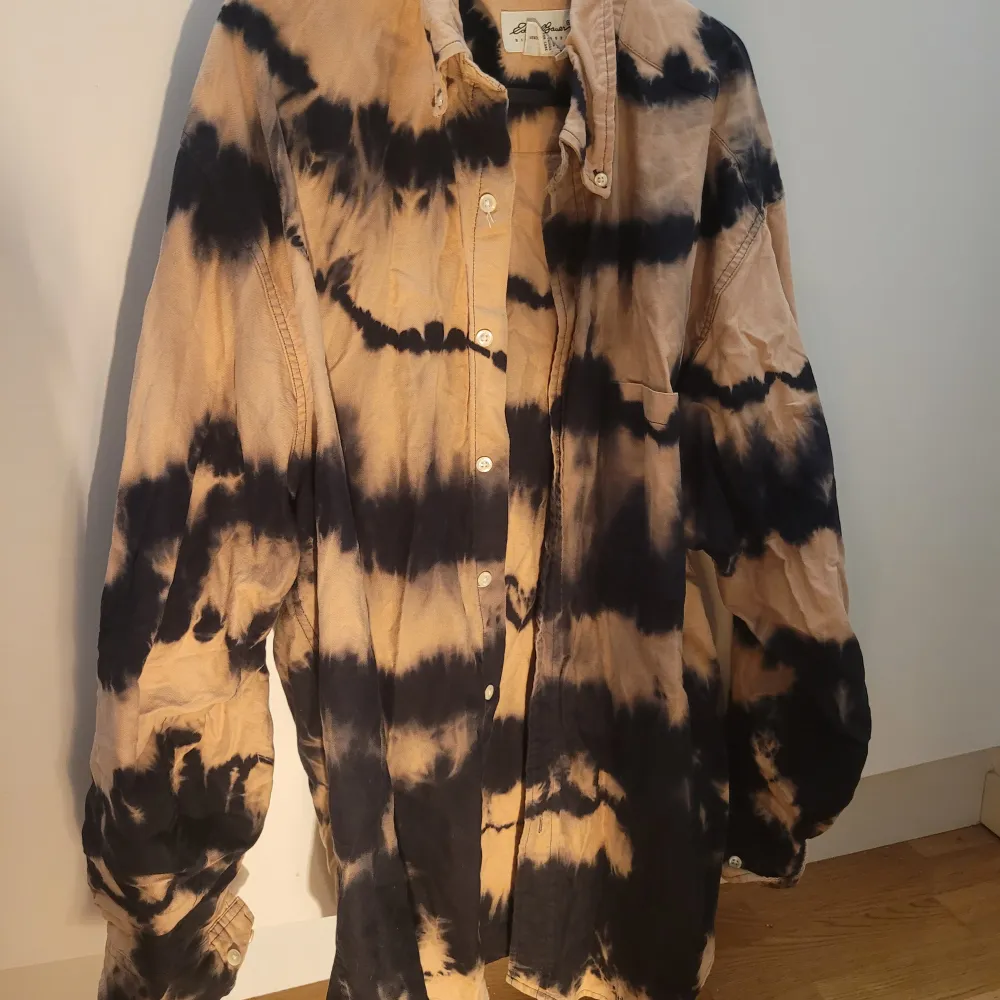 Säljer en unik beige och svart batikmönstrad skjorta med knappar framtill. Skjortan har långa ärmar och en avslappnad passform, perfekt för en cool och avslappnad stil. Den är tillverkad i ett mjukt material som känns skönt mot huden.. Paidat.