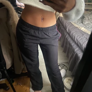 Svarta träningsbyxor från Nike - Säljer ett par svarta träningsbyxor från Nike med rosa detaljer. Byxorna har en elastisk midja och en liten Nike-logga på sidan. Perfekta för träning eller en avslappnad dag. Jätte snygga och skulle säga att de är storlek 34-36.