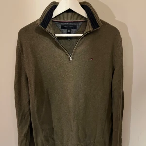 Half zip Olivgrön hoodie från Tommy Hilfiger - Säljer en stilren olivgrön tröja från Tommy Hilfiger med dragkedja vid halsen. Tröjan har långa ärmar och en liten logotyp på bröstet. Perfekt för en avslappnad men snygg look.