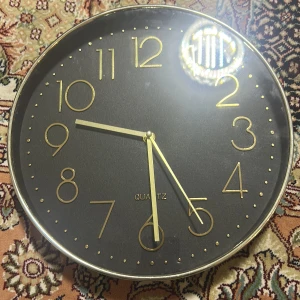 Svart och guld väggklocka - Elegant väggklocka med svart urtavla och guldiga siffror och visare. Klockan har en kvartsrörelse och en stilren design med en tunn, silverfärgad ram. Perfekt för att ge en modern touch till vilket rum som helst.