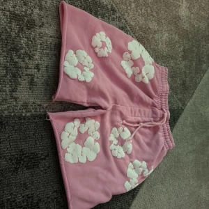 Rosa shorts med vita blommor från denim tears - Säljer ett par rosa shorts från denim tears med vita blommiga mönster. Shortsen har resår i midjan och snörning för extra komfort. Perfekta för varma dagar och har en avslappnad passform.