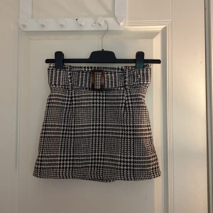 Rutig minikjol med bälte från XOXO Friends - Snygg svartvit rutig minikjol från XOXO Friends med hög midja och dekorativt bälte framtill. Kjolen har klassiskt hundtandsmönster och är perfekt för en trendig look. Med dragkedja på sidan.