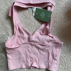 Rosa ribbad linnetopp från H&M - Säljer en ljusrosa ribbad linnetopp från H&M. Toppen har breda axelband och v-ringad halsringning. Tillverkad av återvunnen bomull och har en croppad passform. Perfekt för varma dagar!