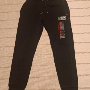Svarta mjukisbyxor från Hoodrich - Svarta mjukisbyxor från Hoodrich med tryckt logga och etikett på ena benet. Byxorna har dragsko i midjan och muddar vid bensluten. Perfekta för en avslappnad stil.