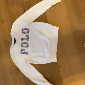 Vit sweatshirt från Polo Ralph Lauren - Vit sweatshirt från Polo Ralph Lauren med färgglada bokstäver på bröstet. Tröjan har rund halsringning och långa ärmar. Perfekt för en avslappnad och stilren look.