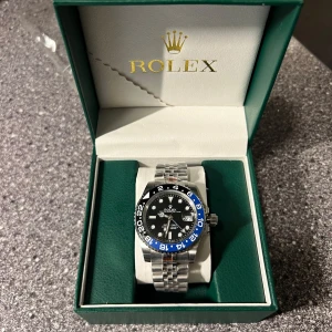 Rolex GMT-Master II “Batman” – Oyster Perpetual, Jubileelänk, Svart/Blå Bezel - Säljer en exklusiv Rolex GMT-Master II med den ikoniska svart/blå “Batman”-bezeln. Klockan har en svart urtavla, datumvisning och 24-timmarsvisare. Levereras i original Rolex-box. Utrustad med jubilee-armband för maximal komfort och elegans.  ✅ Modell: GMT-Master II ✅ Urtavla: Svart med vit text ✅ Bezel: Svart/Blå keramisk ✅ Armband: Jubilee – rostfritt stål ✅ Urverk:  Automatisk ✅ Skick: Nyskick. ✅ Ingår: Originalbox (se bild)