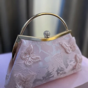 Ljusrosa kuvertväska  - Elegant ljusrosa kuvertväska med guldfärgat metallhandtag och dekorativa fjärilar i broderad spets. Väskan har en glittrig knäppning upptill och en romantisk känsla, perfekt för speciella tillfällen. 