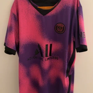 PSG Neymar Jr fotbollströja - Säljer en PSG fotbollströja med Neymar Jr och nummer 10 på ryggen. Tröjan är kortärmad och har en unik lila och rosa färgkombination med svarta detaljer och klubbmärke på bröstet. Perfekt för fans av Neymar och PSG. Som ni ser på baksidan så ”pillas” nån bokstav av (tröjan köptes så i second hand butik) och är använd ett par gånger. Säljer den pga att tröjan passar inte  min stil längre. 