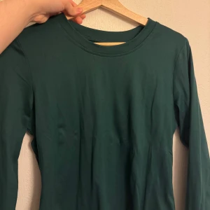 Mörkgrön långärmad topp - En enkel och stilren mörkgrön långärmad topp med rund halsringning. Perfekt att matcha med jeans eller kjol för en clean look. Mjuk och skön mot huden. I storlek L men liten i storlek