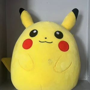Pikachu Squishmallow - En supermjuk och kramvänlig Pikachu Squishmallow! Perfekt för Pokémon-fans som vill ha en mysig kompis i soffan eller sängen. Den ikoniska gula färgen och de röda kinderna gör den till ett måste för alla unga samlare. Tvättas såklart före leverans!