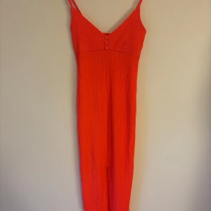 Orange långklänning från Gina Tricot - Säljer en snygg orange långklänning från Gina Tricot med smala axelband och v-ringning. Klänningen har dekorativa knappar framtill och är gjord i ett strukturerat, syntetiskt material. Perfekt för sommarens alla tillfällen!