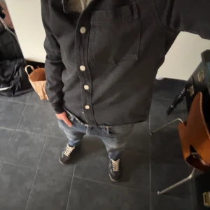 Svart overshirt med knappar - Säljer en svart overshirt med klassisk krage och vita knappar framtill. Jackan har en bröstficka på vänster sida och är långärmad. Perfekt att slänga över en hoodie eller t-shirt för en clean look.