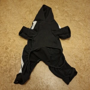 Svart hoodie för hundar - Svart hoodie med huva och vita ränder längs ärmar och ben. Tröjan har lång ärm och är helsvart förutom de vita detaljerna. Perfekt för en sportig look.