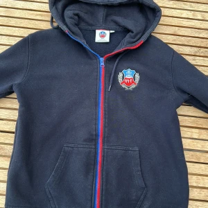 Hoodie från Helsingborgs IF - Svart hoodie med huva och broderat HIF-logga på bröstet. Tröjan har en dragkedja i rött och blått samt två stora fickor framtill. Perfekt för dig som vill visa ditt stöd för HIF.
