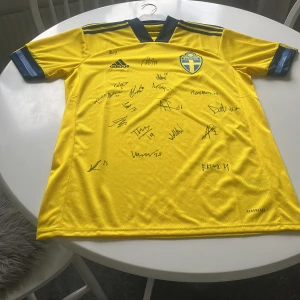 Svenska herrlandslagets fotbollströja från Adidas - Gul kortärmad fotbollströja från Adidas med blå detaljer och svenska landslagets emblem på bröstet. Tröjan har svarta ränder på axlarna och är tillverkad i lätt, ventilerande material (AEROREADY). Flera autografer på framsidan som till exempel Williot swedberg och Hugo Larsson samt andra lanslasspelare.Skicket på tröjan är 10/10 eftersom att den aldrig är använd! Lägg pris förslag!