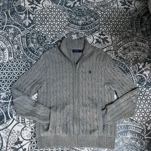 Grå kabelstickad kofta från Ralph Lauren - Säljer en grå stickad kofta från Polo Ralph Lauren med dragkedja framtill. Tröjan har kabelstickat mönster, hög krage och ribbade muddar vid ärmslut och nederkant. Klassisk liten logga på bröstet.