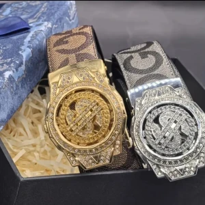 Bälte med glittrande spänne och GG-mönster - Säljer ett bälte med stort, glittrande spänne i guld eller silverfärg. Bältet har tydligt GG-mönster på bandet i bruna eller grå toner. Spännet är dekorerat med stenar och har en unik design med dollartecken.