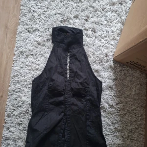 Svart ärmlös halterneck topp - Säljer en svart ärmlös halterneck topp med hög krage och knappdetaljer framtill. Toppen har en snygg öppning framtill och markerade sömmar som ger en cool look.