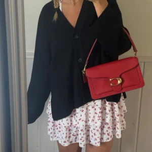 Röd axelväska från Coach - Säljer en snygg röd axelväska från Coach i skinn med guldfärgade metalldetaljer och ett stort C-spänne framtill. Väskan har flera fack inuti och en justerbar axelrem. Perfekt för att lyfta vilken outfit som helst!