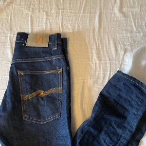Mörkblå jeans från Nudie Jeans - Snygga mörkblå jeans från Nudie Jeans med klassiska orange sömmar och femficksdesign. Byxorna har normal passform och raka ben, samt detaljer som dragkedja och knapp i midjan. Perfekta för en stilren look.