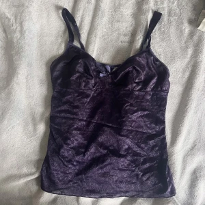 Lila spetslinne med rosett - Säljer en lila linnetopp i spets med smala axelband och en liten rosett framtill. Toppen har en lätt glansig yta och figurnära passform, perfekt att styla till olika outfits. Den är även lite genomskinlig. Storlek : 38-40, M