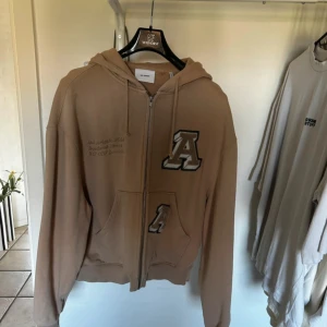 Beige hoodie med dragkedja från Axel Arigato - Säljer en beige hoodie från Axel Arigato med dragkedja framtill och huva. Tröjan har två fickor och stora A-tryck på bröstet och fickan. Perfekt för en avslappnad stil.