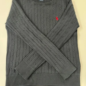 Svart kabelstickad tröja från Ralph Lauren - Svart kabelstickad tröja från Ralph Lauren med rund halsringning och röd broderad logga på bröstet. Tröjan har långa ärmar och ribbade muddar vid ärmslut och nederkant. Klassisk och stilren modell. Perfekt för våren och kallare dagar