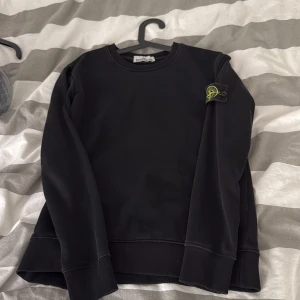 Svart sweatshirt från Stone Island - Säljer en svart sweatshirt från Stone Island med klassisk logotyp-patch på vänster ärm. Tröjan har rund halsringning och ribbade muddar vid ärmslut och nederkant. Perfekt för en stilren och avslappnad look. Den är äkta men inget sätt att bevisa de på därav de låga priset 