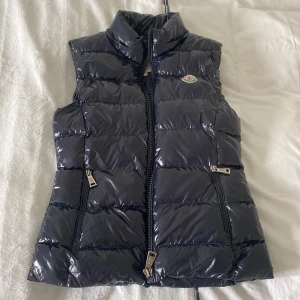 Glansig dunväst från Moncler - Snygg svart dunväst från Moncler med hög krage och två dragkedjefickor framtill. Västen har en glansig finish och klassisk Moncler-logga på bröstet. Modellen är figursydd och går in i midjan. Storleken motsvarar XS🤗